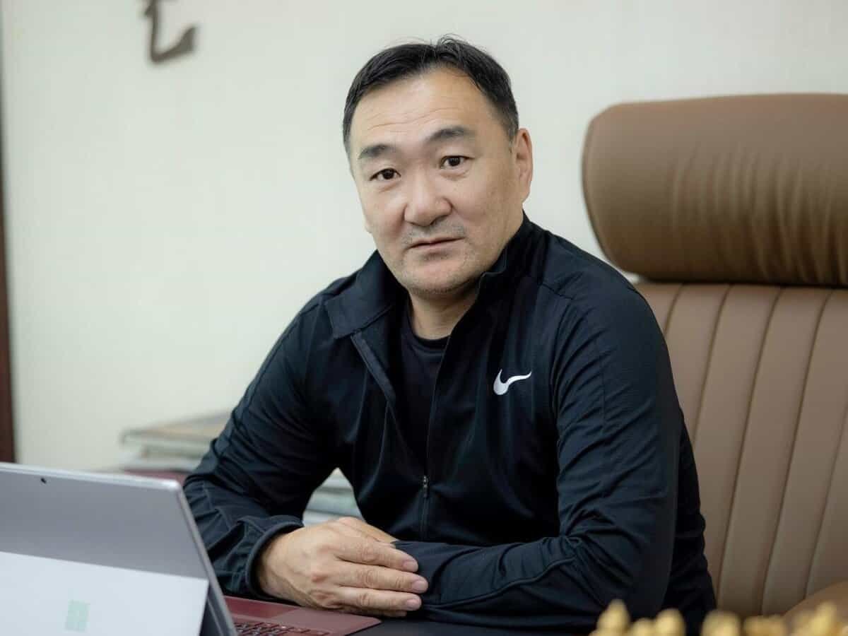 Богино хар үстэй хүн ширээний ард сууж байна. Тэр хар Nike хүрэм өмсөж, камер руу харж байна. Түүний өмнө зөөврийн компьютер байгаа нь бизнесээ удирдаж буй энтрепренер байж магадгүй юм. Түүний ард хүрэн өндөр түшлэгтэй сандал байгаа бол арын дэвсгэр нь цайвар, бүдгэрсэн хэвээр байна. - Business.MN