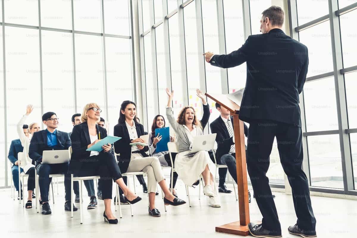 Бизнесийн мэргэжилтэн, энтрепренер эгнээнд суусан үзэгчдэд илтгэл тавьж байна. Зарим гишүүд том цонхноос байгалийн гэрлээр дүүрсэн орчин үеийн хурлын танхимд гараа өргөж байна. - Business.MN