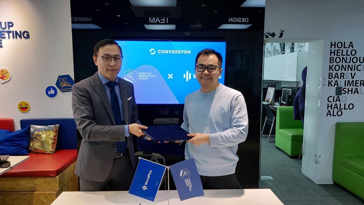 Оффисын орчинд хоёр эрэгтэй гар барьж байна. Нэг нь саарал костюм, зангиа өмссөн энтрепренер, нөгөө нь цайвар цэнхэр цамц, нүдний шилтэй. Тэд цэнхэр хавтас барьж байна. Арын дэвсгэр дээр "CONVERYSON" болон бусад бизнесийн лого бүхий том дэлгэц харагдана. - Business.MN