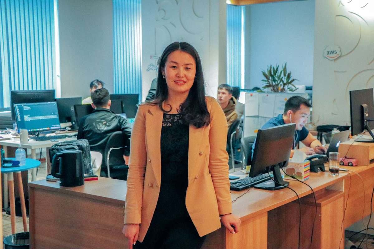 Энтрепренер бололтой шаргал пиджак өмссөн эмэгтэй завгүй оффисын ширээний хажууд компьютер дээр ажилладаг хүмүүстэй инээж байна. - Business.MN