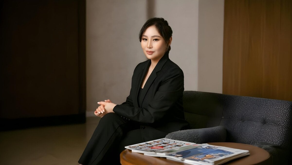 A person in a black suit sits on a dark sofa, hands clasped, with a neutral expression. A table in the foreground has magazines placed on it. The background is softly lit with muted colors.
