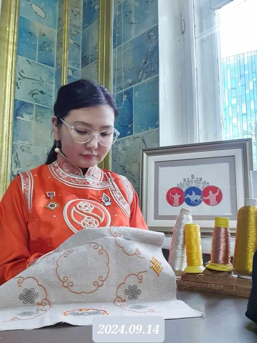 A person in an orange traditional outfit is engaged in embroidery work. They sit by a window with blue tiles and a framed design. Several spools of thread are on the table. The date "2024.09.14" is overlaid at the bottom.