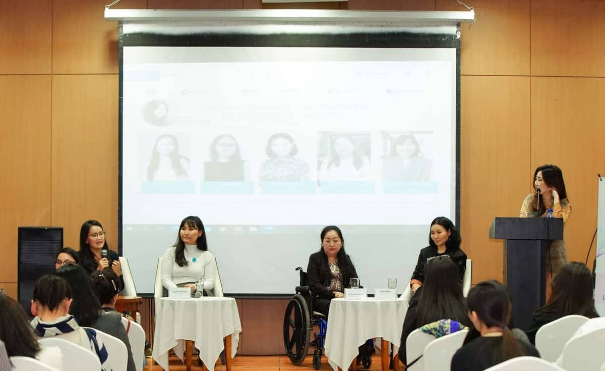 Таван бизнес эрхлэгч эмэгтэйчүүдийн нэг нь тэргэнцэртэй, ард түмнийхээ өмнө гэрэл зургуудыг нь харуулсан дэлгэцээр бизнесийн ертөнцөөр аялах тухай ойлголтоо хуваалцаж, үзэгчдийн өмнө ярилцаж байна. - Business.MN