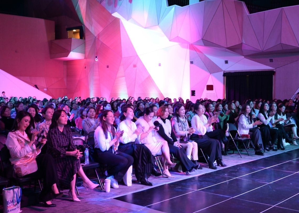A large audience sits in rows, attentively watching a performance or presentation. The space features modern, angular architecture with colorful lighting in shades of pink and purple. Many in the audience are clapping, creating a lively atmosphere.