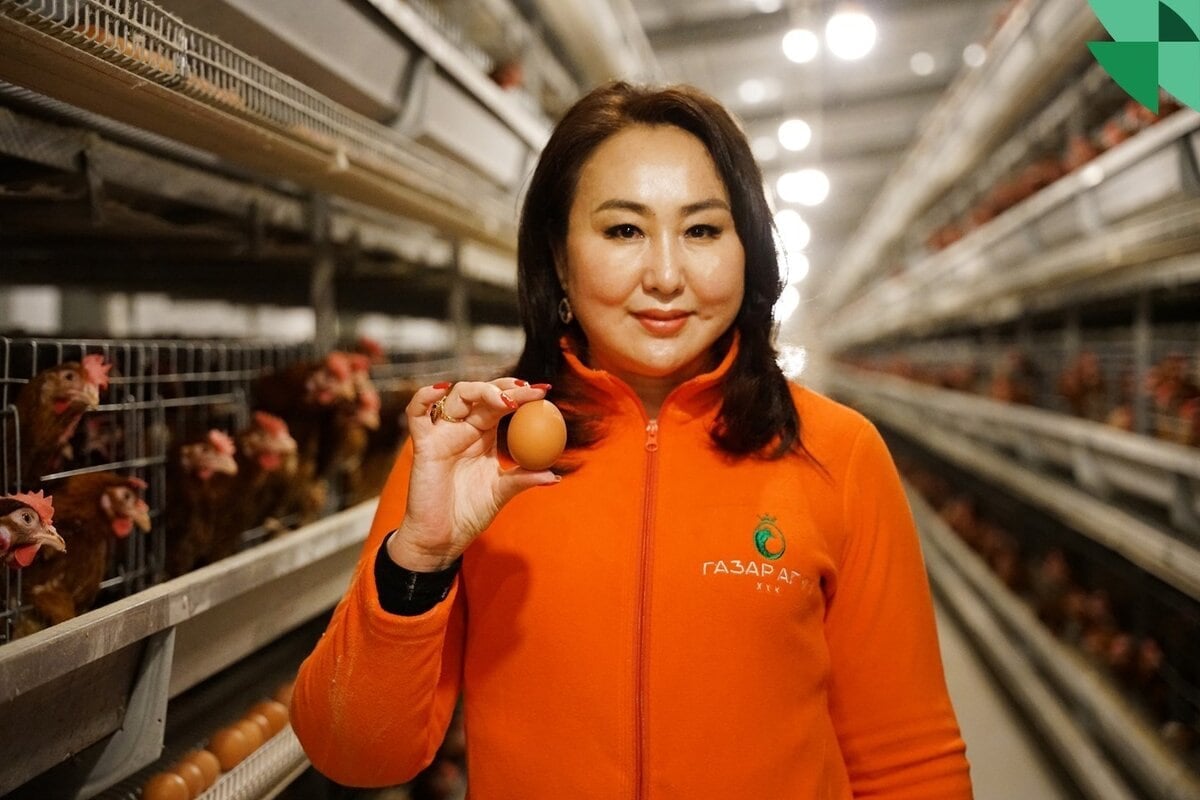 A woman in an orange jacket stands in a poultry farm aisle, holding an egg. Chickens are in cages on both sides. The setting is well-lit, and she appears confident and focused.