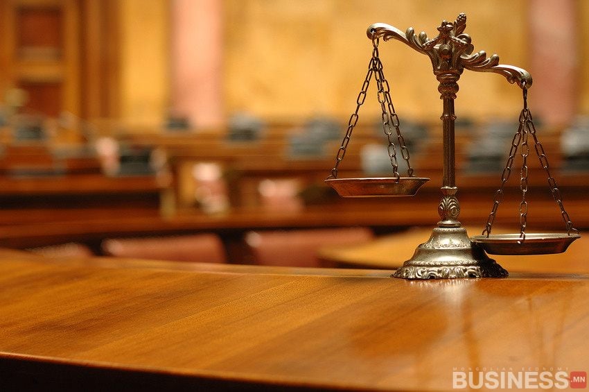 Шүүхийн хоосон танхимд модон ширээн дээр шударга ёсны хүрэл жинлүүр суусан нь ямар ч бизнест нөлөөлж болох хүнд шийдвэрүүдийг тусгаж, цаана нь бүдгэрсэн суудалтай. - Business.MN