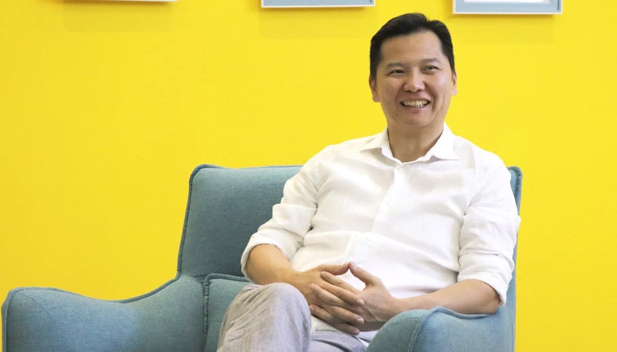 Цагаан цамцтай эр тод шар хананд наалдсан цэнхэр сандал дээр тухтай сууж байна. Тэр инээмсэглэж, тайвширч, гараа өвөр дээрээ атгасан мэт харагдаж байна. - Business.MN