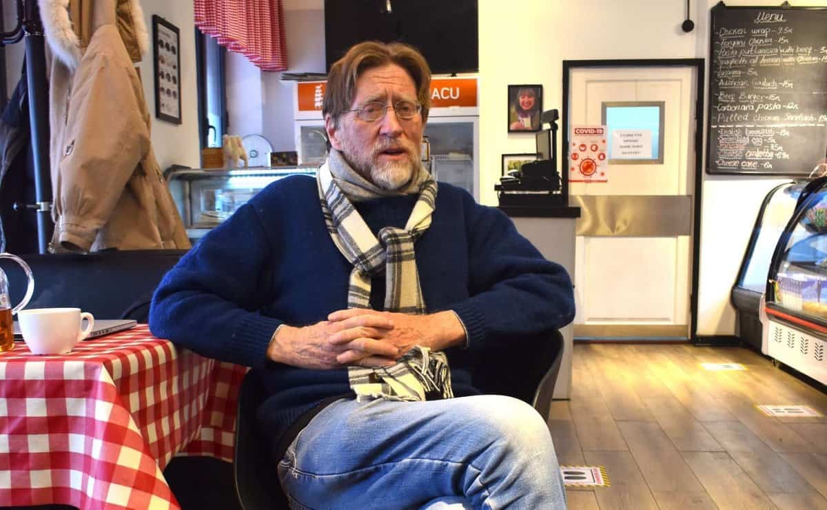 Нүдний шил, ороолттой эр цэнхэр цамц, жинсэн өмд өмссөн кафед сууж байна. Кафе нь алаг ширээний бүтээлэг, өлгөөтэй пальто, ханан дээр цэсний самбартай. Түүний хажууд ширээн дээр аяга байна. - Business.MN