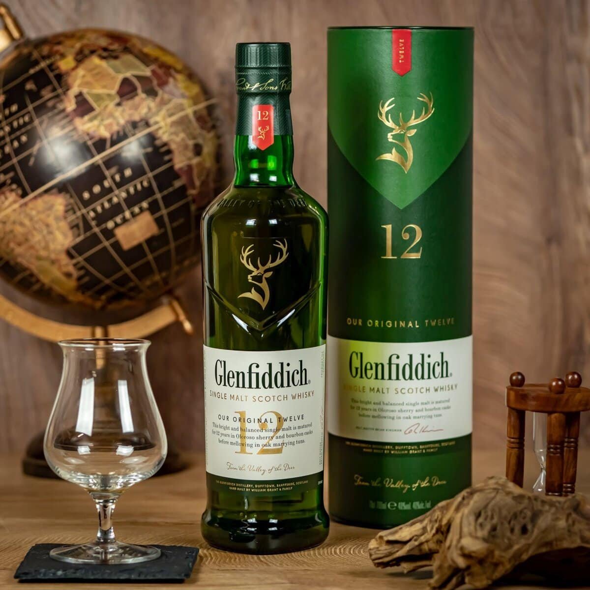 A bottle of Glenfiddich 12-year-old single malt Scotch whisky on a wooden table, accompanied by its cylindrical box. A globe, an empty whisky glass on a coaster, and a small wooden chair are nearby, creating a cozy, sophisticated setting.