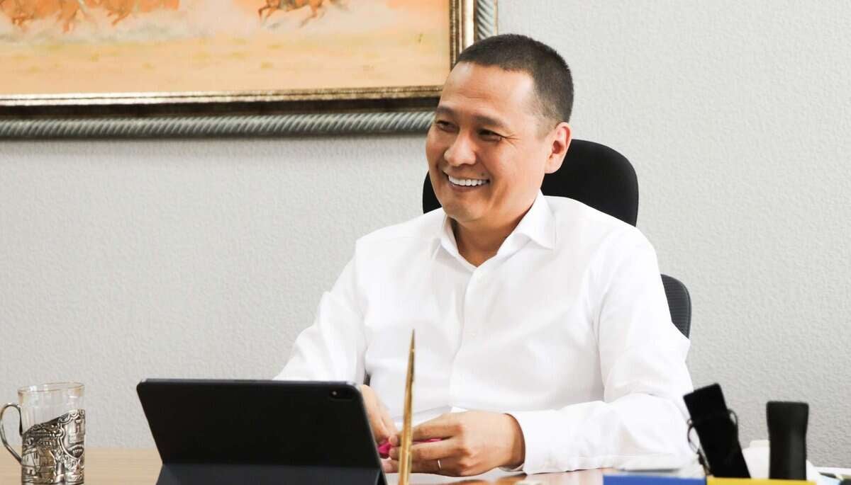 Цагаан цамц өмссөн инээмсэглэсэн энтрепренер ширээний ард таблет, цаас барин суугаад дараагийн бизнесээ төлөвлөж байна. Арын ханан дээр зураг байна. - Business.MN