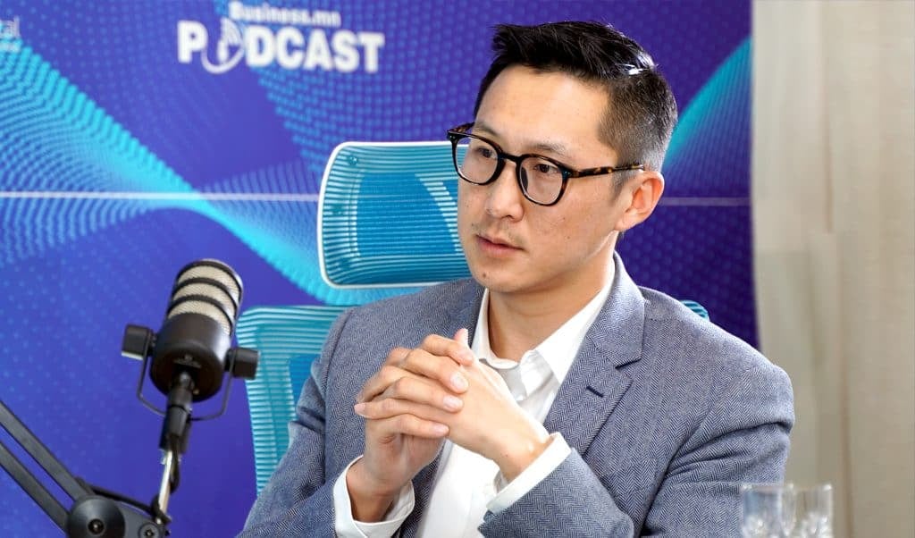 Нүдний шил, цагаан цамц, цэнхэр пиджак өмссөн хүн микрофонд суугаад ярьж байна. Тэдний ард дууны долгионы хээ болон "PODCAST" гэсэн үг бүхий арын дэвсгэр байдаг. Энтрепренер яриа эсвэл ярилцлагад оролцож байгаа бололтой. - Business.MN