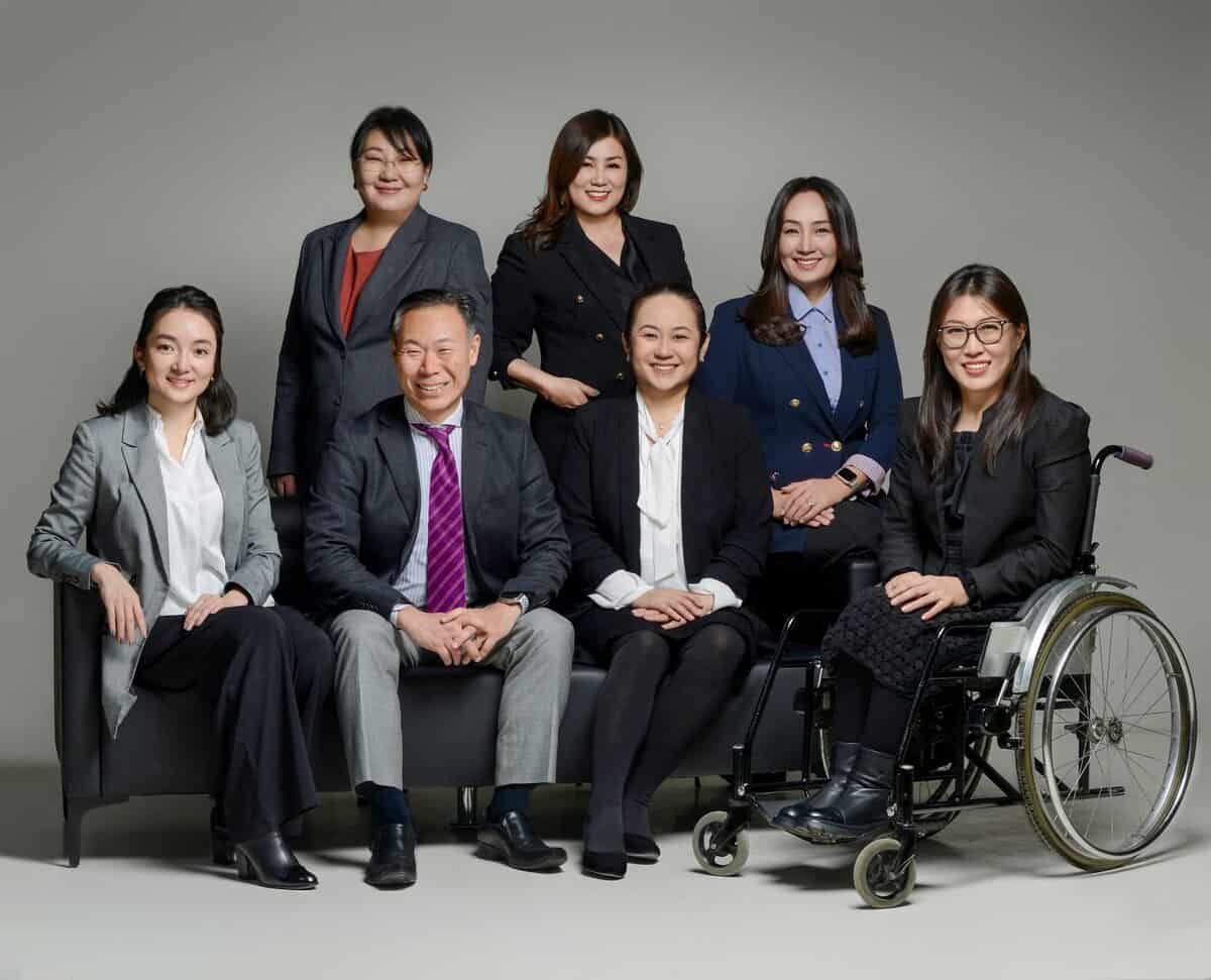 Дөрвөн эмэгтэй зогсож, гурван хүн сууж буй бизнесийн долоон мэргэжилтнүүдийн нэг нь нил ягаан өнгийн зангиа зүүсэн эрэгтэй, нөгөө нь тэргэнцэртэй энтрепренер бөгөөд бүгд камерын өмнө харан, саармаг саарал дэвсгэртэй студийн орчинд инээмсэглэж байна. - Business.MN