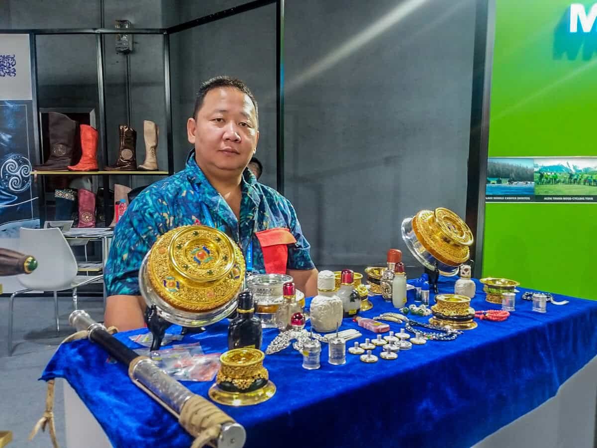 Нэг хүн уламжлалт эд өлгийн зүйлс, тэр дундаа гоёл чимэглэлийн алтан эдлэл болон бусад жижиг эд зүйлсийг харуулсан ширээний ард сууж байна. Ширээ нь цэнхэр даавуугаар хучигдсан байдаг. Цаана нь дэлгэсэн эд зүйлс бүхий тавиурууд, бичвэр бүхий ногоон хана байдаг. - Business.MN