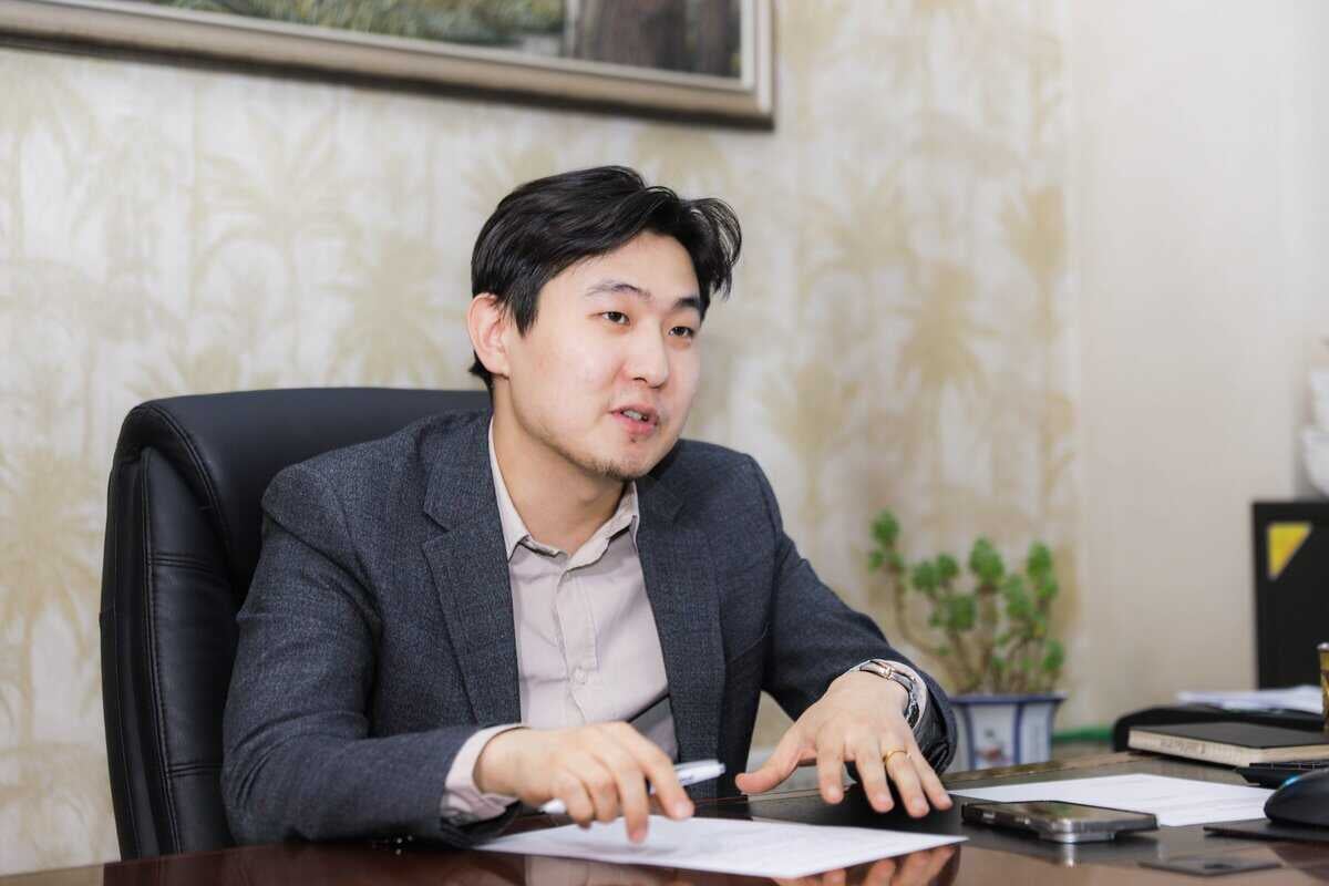 A man in a gray suit is seated at a desk with papers. He's gesturing with one hand while speaking. On the desk are a pen, a notebook, and a smartphone. In the background, there's a framed picture and a potted plant.