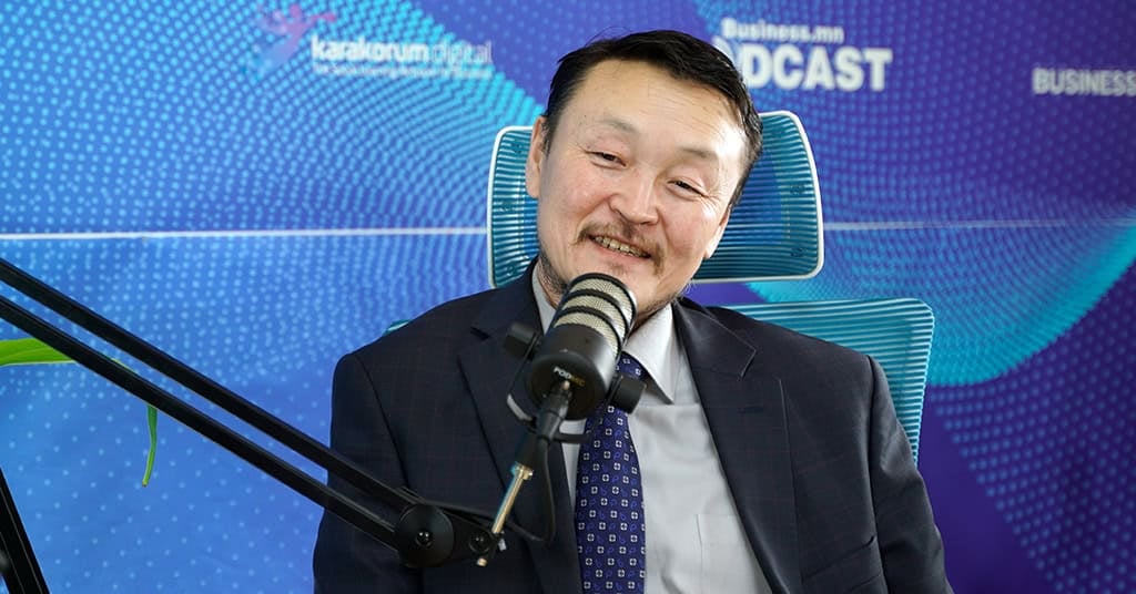 Хар хөх алаг хослол, цэнхэр тасархай зангиа өмссөн хүн микрофонд ярьж байна. Тэрбээр "Каракорум дижитал", "Business.mn Подкаст" гэсэн бичвэр бүхий цэнхэр дэвсгэрийн өмнө сууж, энтрепренерийн аяллаа ярилцаж байна. - Business.MN