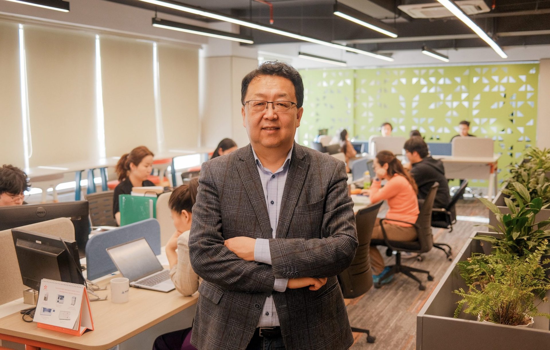 A man in a checked blazer stands with arms crossed in a modern office, smiling at the camera. Several people work on laptops at desks in the background. The space has green decor, plants, and large windows.