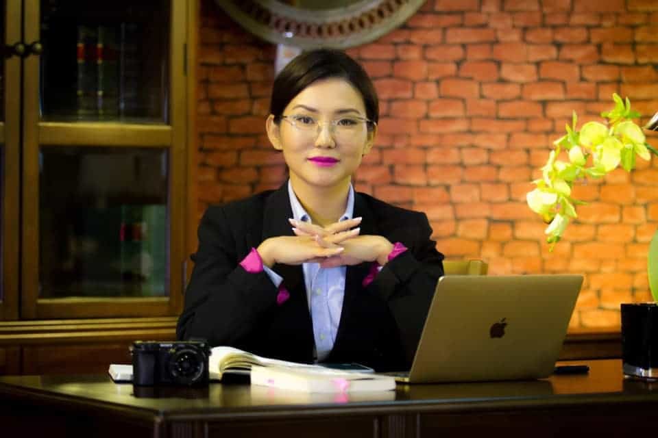 Нүдний шил зүүсэн эмэгтэй ширээний ард суугаад зөөврийн компьютер, камер, номтой, тавиур бүхий тоосгон хананд хүрээтэй байна. Энэхүү энтрепренер нь компанийхаа дижитал ландшафтыг чиглүүлэхдээ бизнесийн сүнсийг шингээдэг. - Business.MN