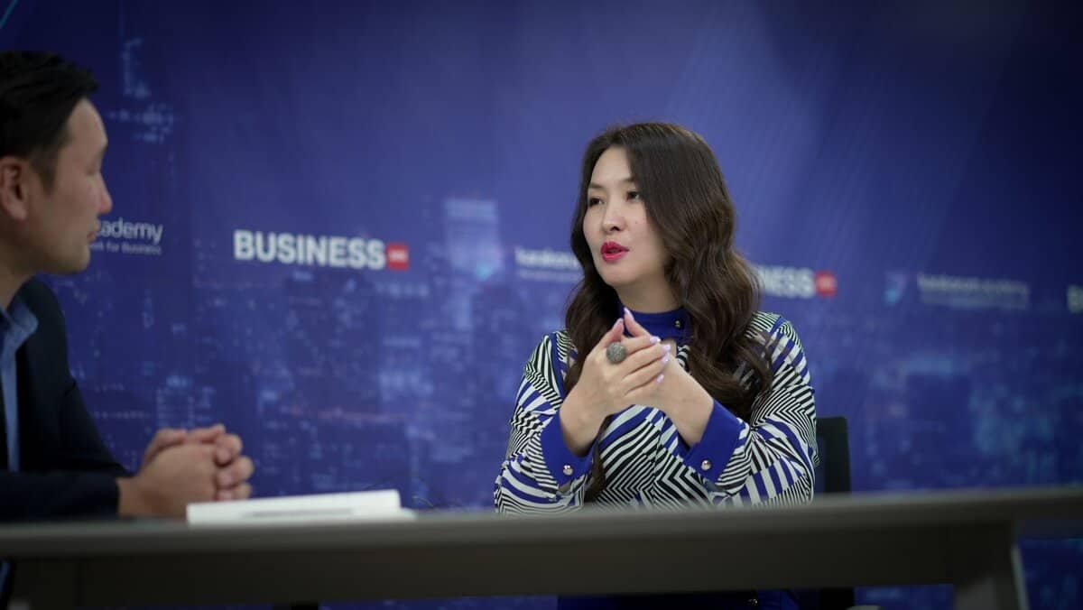 Урт үстэй, хээтэй цамц өмссөн эмэгтэй энтрепренертэй ярилцаж байхдаа гараараа дохиж ярьж байна. Тэд бизнес, эрдэм шинжилгээний холбогдолтой бичвэр бүхий цэнхэр дэвсгэрийн эсрэг ширээний ард сууж байна. - Business.MN