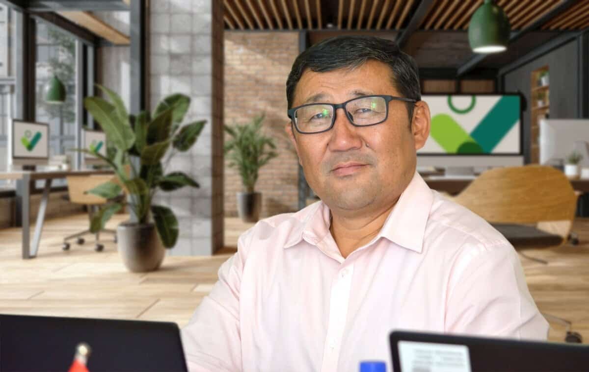 Нүдний шил, цайвар ягаан цамц өмссөн эрэгтэй ширээний ард сууж, урд нь зөөврийн компьютер байна. Ар талд нь орчин үеийн оффисын доторх ургамлууд, модон шал, тоосгон ханан дээрх том дэлгэц бүхий шинэлэг бизнесийн орчны уур амьсгалыг төгс тусгасан болно. - Business.MN