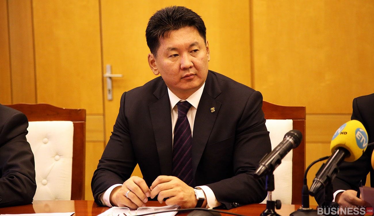 Костюм өмссөн хүн урд нь микрофонтой ширээний ард сууна. Тэрээр албан ёсны уулзалт эсвэл хэвлэлийн бага хурал дээр байгаа бололтой. Арын дэвсгэр дээр модон хана, хаалга харагдана. - Business.MN