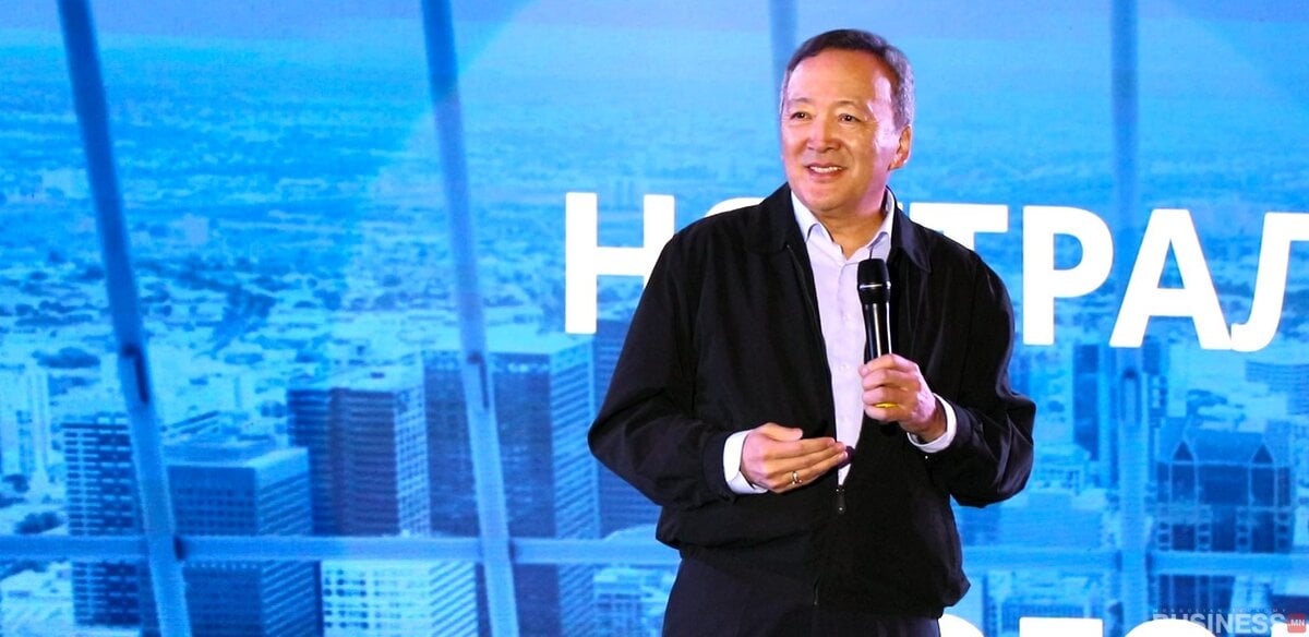 Харанхуй хүрэмтэй энтрепренер тайзан дээр микрофоноор ярьж байна, ард нь хотын өнгө үзэмж харагдана. - Business.MN