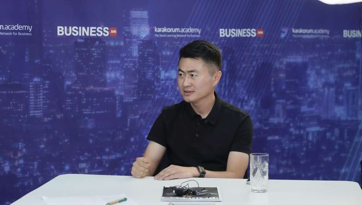 Хар цамцтай хүн ширээний ард дэвтэр, аяга устай урдаа суугаад ярьж байна. Арын дэвсгэр дээр шөнийн цагаар хотын өнгө үзэмж, "BUSINESS" болон "karakoum.academy" гэсэн логонууд байгаа нь түүнийг хамгийн сүүлийн үеийн компанийхаа талаар ярилцаж буй энтрепренер гэдгийг харуулж байна. - Business.MN