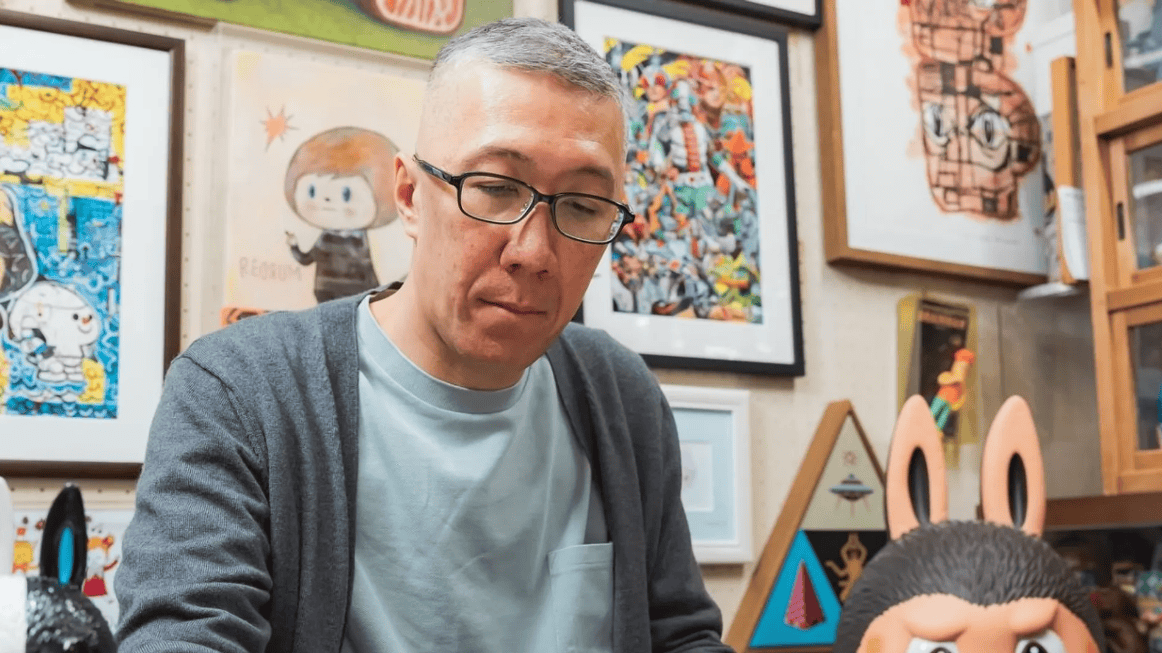 Богино буурал үстэй, нүдний шилтэй хүн ширээний ард ажиллаж байхдаа саарал кардиган, цэнхэр цамц өмсөж, өнгө өнгийн урлагийн бүтээлүүд, жаазтай зургууд, ханан дээр дүрсэлсэн, хөгжилтэй Лабубу үлгэрийн баатраар хүрээлэгдсэн байдаг. - Business.MN