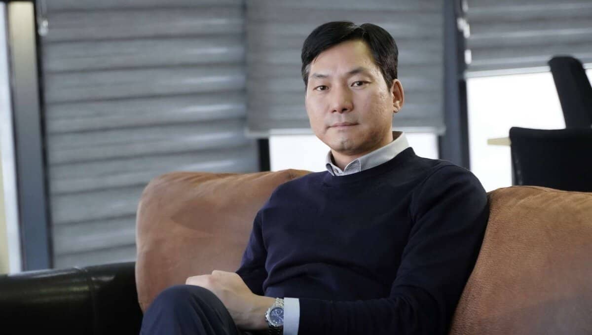 Богино хар үстэй, цагаан захтай цамцан дээр хар хөх өнгийн ноосон цамц өмссөн эрэгтэй бор буйдан дээр сууж байна. Төвийг сахисан илэрхийлэлтэйгээр, арын хэсэгт металл цонхны наалт бүхий дотоод орчинд тунгалаг харагдаж байна. - Business.MN