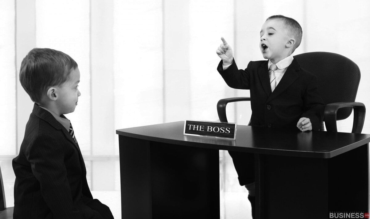 Костюм өмссөн хоёр залуу оффисын орчинд байна. "Дарга" гэсэн шошготой ширээний ард сууж буй нэг хүү заагаад хөдөлгөөнтэй ярьж байна. Нөгөө хүү түүний эсрэг суугаад анхааралтай сонсож байна. Энэ дүр зураг хар цагаан өнгөтэй байна. - Business.MN