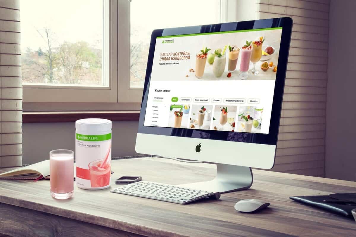 Модон ширээн дээр смүүтиний жор бүхий вэбсайтыг харуулсан ширээний компьютер сууж байна. Хажууд нь Herbalife коктейль хольцтой сав, ягаан коктейлээр дүүргэсэн шил байна. Нарны гэрэл гадаа мод харагдах цонхоор шүүнэ. - Business.MN