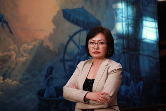 Харанхуй, бүрхэг өнгө бүхий том зургийн өмнө нүдний шилтэй, гараа зөрүүлсэн эмэгтэй зогсож байгаа нь бизнесийн ертөнцийг байлдан дагуулахад бэлэн байгаа өөртөө итгэлтэй энтрепренерийн дүр төрхийг илэрхийлдэг. - Business.MN