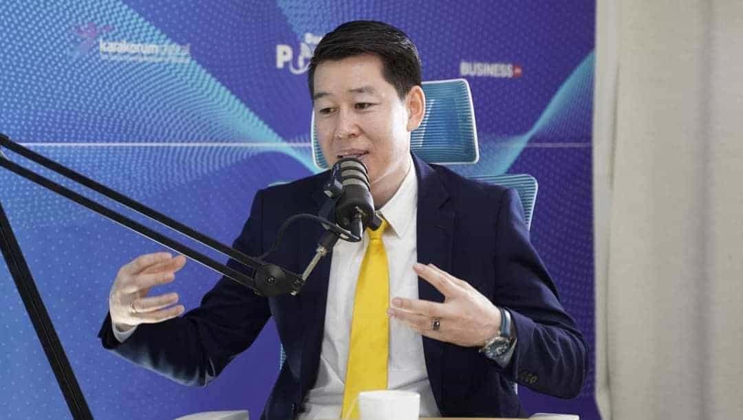 Хар өнгийн костюм өмссөн шар зангиа зүүсэн эр микрофоны өмнө суугаад ярьж, гараараа дохиж байна. Тэрээр цэнхэр оффисын сандал дээр сууж, цэнхэр, нил ягаан өнгийн дэвсгэр дээр бизнесийн тухай бичвэр, хэв маягийг харуулсан. Түүний урд ширээн дээр цагаан аяга байна. - Business.MN