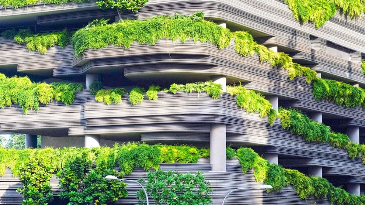 A modern building with tiered, curving balconies covered in lush green plants and foliage, creating a vertical garden effect, with sunlight highlighting the vibrant greenery.