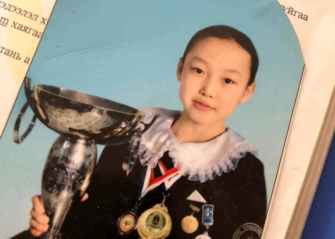 Сургуулийн дүрэмт хувцастай, медаль, энгэрийн тэмдэгтэй залуу цайвар цэнхэр дэвсгэр дээр сууж буй том мөнгөн цомыг барьж байна. Гэрэл зургийн хажууд Нарны цирк, Б.Одгэрэл нарын Сөүл дэх ололт амжилтыг харуулсан баримт бичиг хэсэгчлэн харагдаж байна. - Business.MN