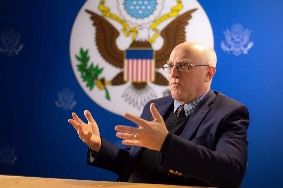 Ширээний ард сууж байхдаа нүдний шил, бараан костюм өмссөн, зангиа зүүсэн халзан эр гараараа дохио зангаа. Түүний ард халзан бүргэд дүрсэлсэн АНУ-ын том сүлд байдаг бөгөөд хөх дэвсгэр дээр бэлгэдлийн жижиг хувилбаруудыг харуулсан бөгөөд энэ нь түүний нөлөө бүхий бизнес эрхлэгчийн үүргийг бэлгэддэг. - Business.MN