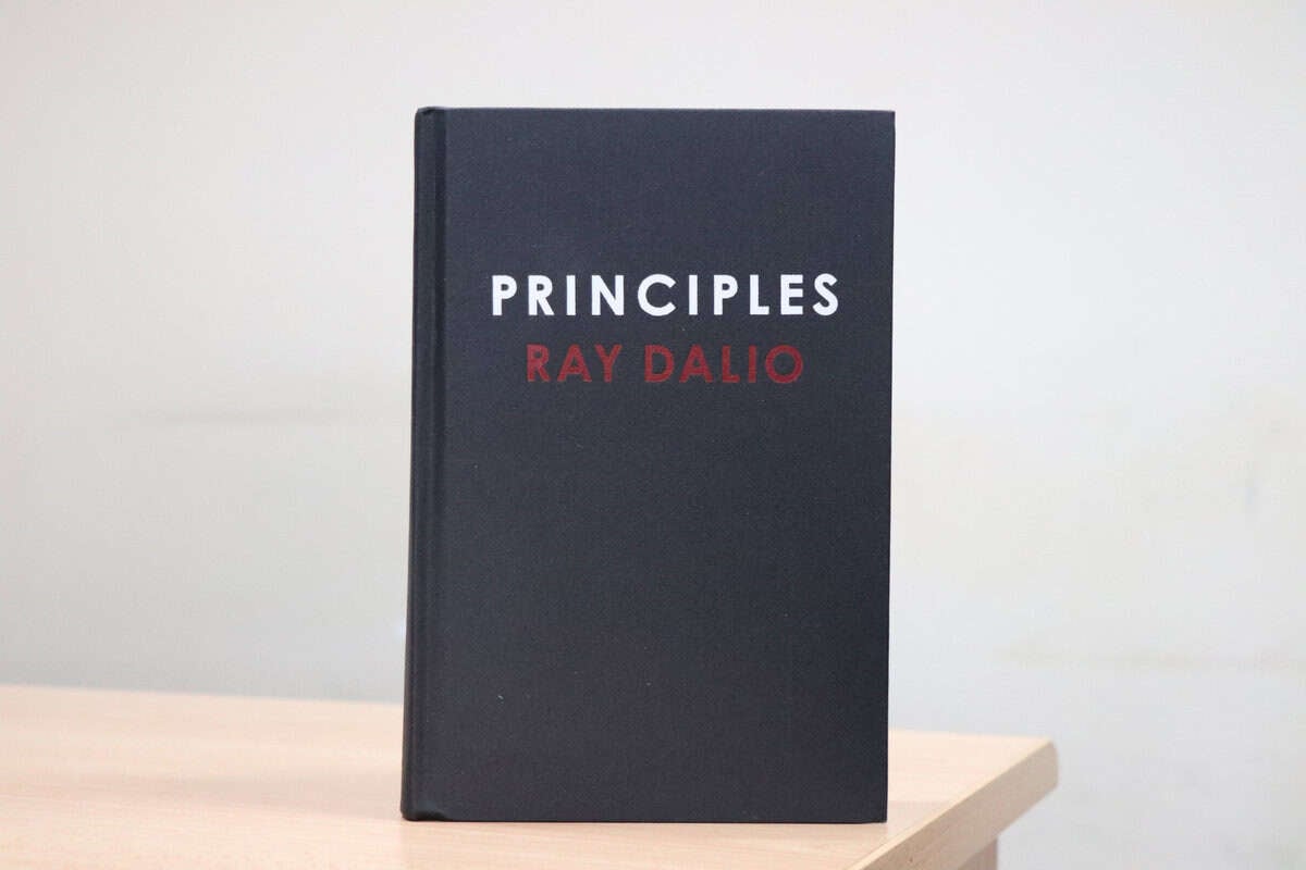 Рэй Далиогийн "Зарчмууд" нэртэй хатуу хавтастай ном цайвар модон гадаргуу дээр босоо зогсож байна. Хавтас нь хар өнгөтэй, гарчиг нь цагаан, зохиогчийн нэр улаан өнгөтэй байна. Арын дэвсгэр нь энгийн, цайвар өнгөтэй. - Business.MN