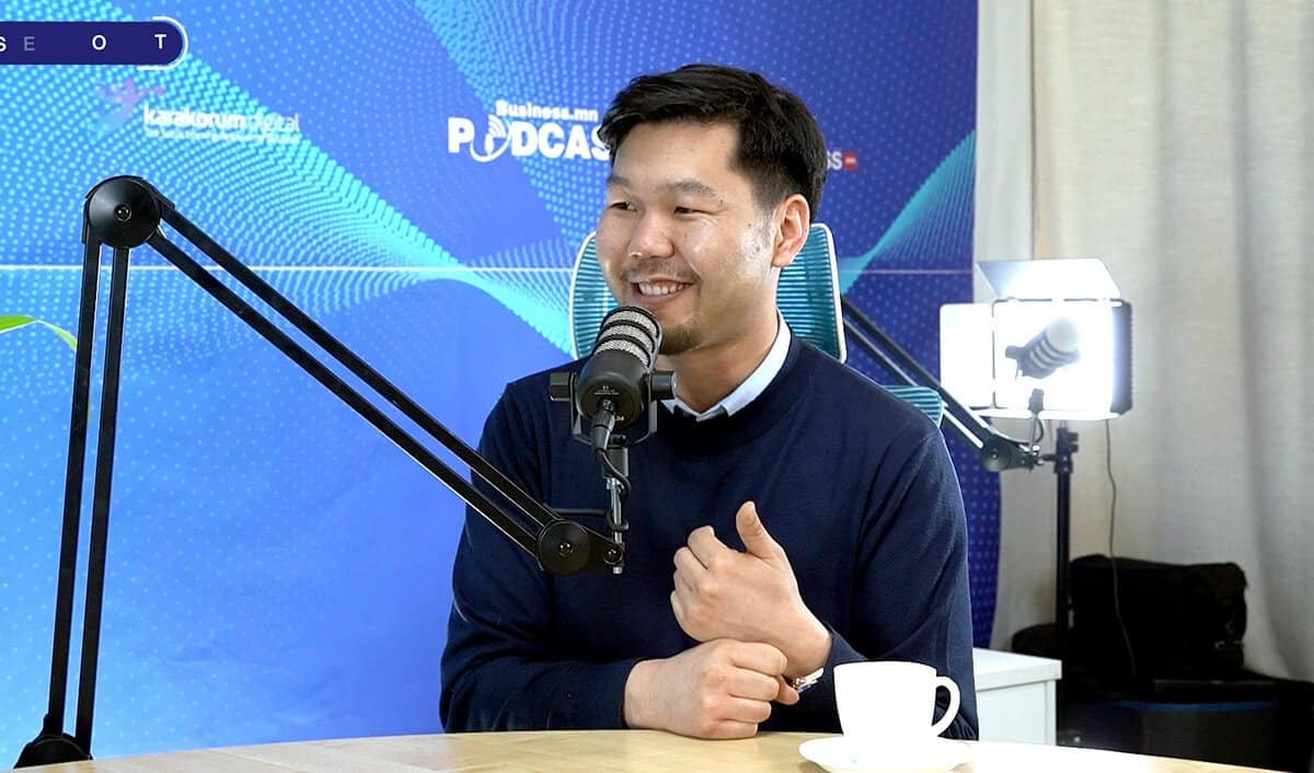 Харанхуй ноосон цамц өмссөн эр аяга кофе барин ширээний ард суугаад микрофоноор ярьж байна. Түүний ард подкастыг сурталчилсан бичвэр бүхий цэнхэр дэвсгэр байна. Тэр энтрепренер бололтой, яриа өрнүүлж, инээмсэглэж, гараараа дохиж байна. - Business.MN