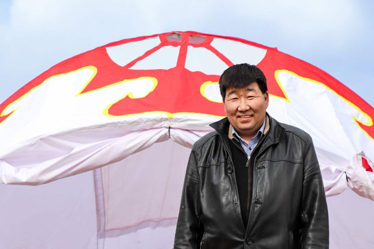 Хар савхин хүрэм өмссөн хүн цаанаа улаан цагаан өнгийн том байшингийн өмнө инээмсэглэж байна. Тэнгэр багавтар үүлтэй. - Business.MN