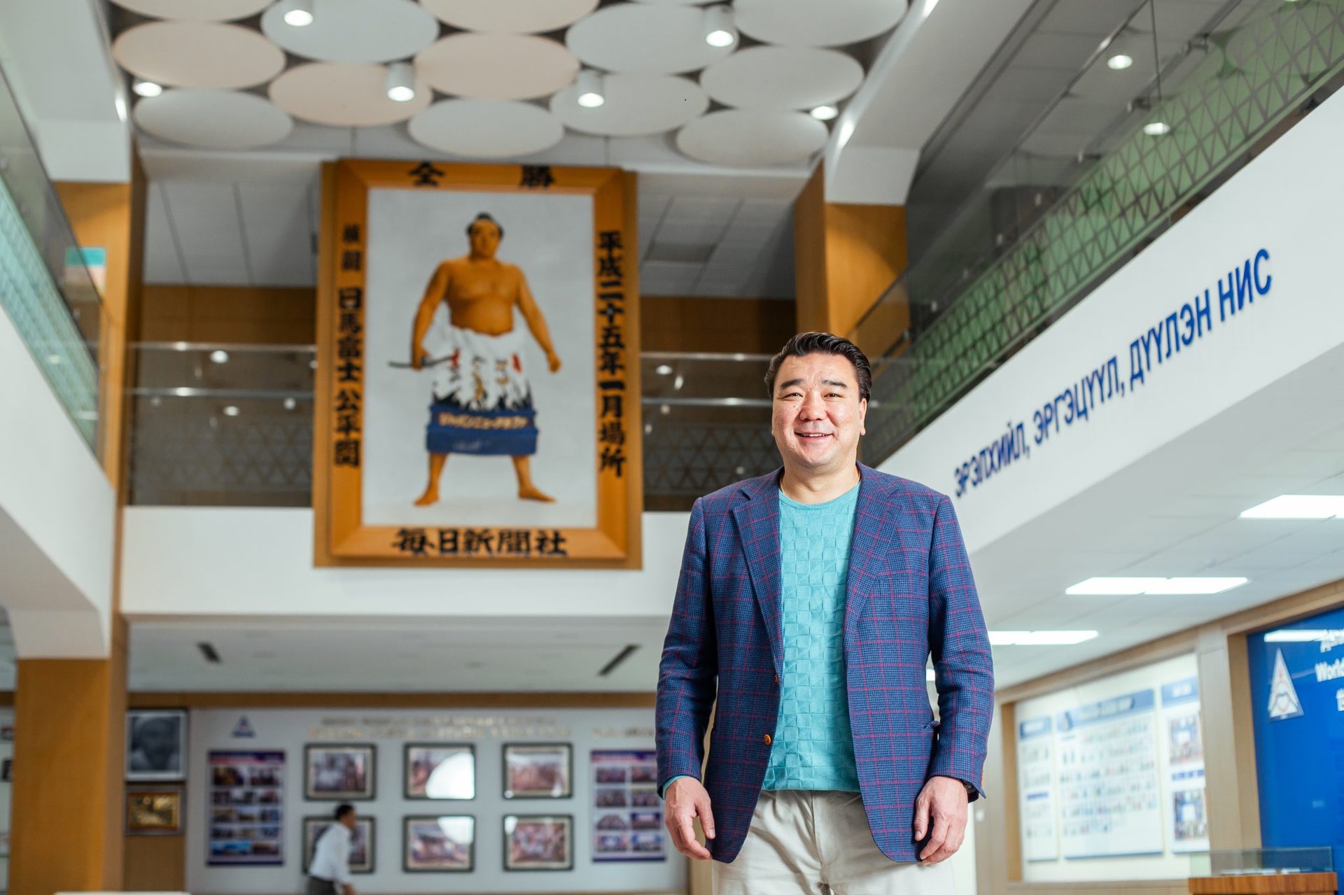 A man in a blue patterned blazer and jeans stands smiling in a building lobby. Behind him is a large portrait of a sumo wrestler. The walls have signs with text and framed photographs. The ceiling features circular designs.