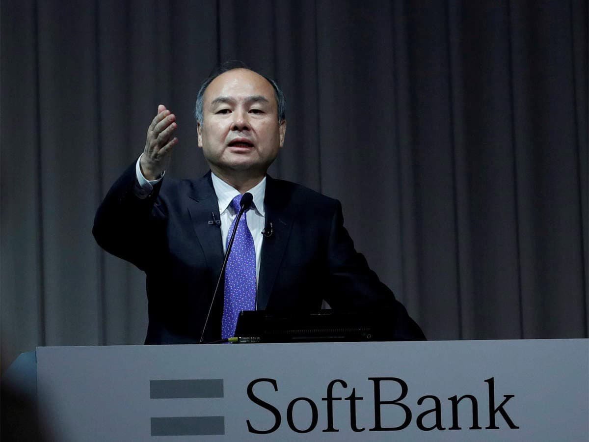 Костюм, зангиа зүүсэн эр SoftBank лого бүхий индэрийн ард зогсож, зүүн гараараа дохиж байна. Арын дэвсгэр нь доторх орчин, магадгүй бага хурал эсвэл хэвлэлийн арга хэмжээг илэрхийлсэн хөшигтэй. Энэхүү энтрепренер нь компанийн бизнесийн гол стратегиудыг итгэлтэйгээр авч үздэг. - Business.MN