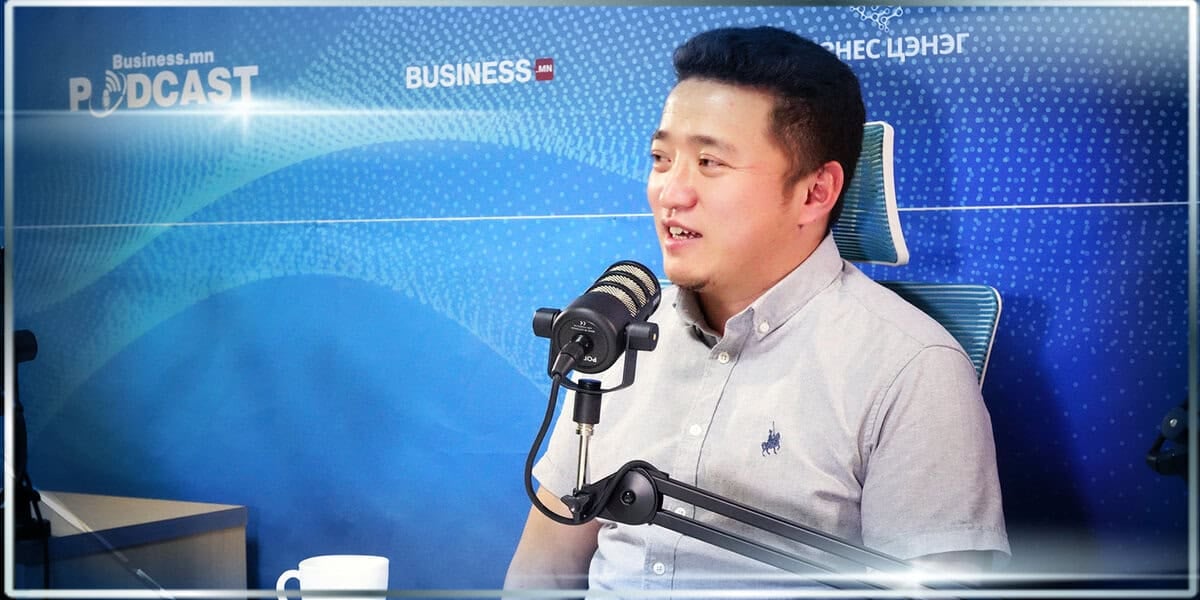Цэнхэр сэдэвт чимэглэл бүхий подкаст бичлэгийн студид микрофон руу товчтой цамцтай эр ярьж, бизнесийн стратеги болон компанийн өсөлтийн талаар ярилцаж байна. - Business.MN