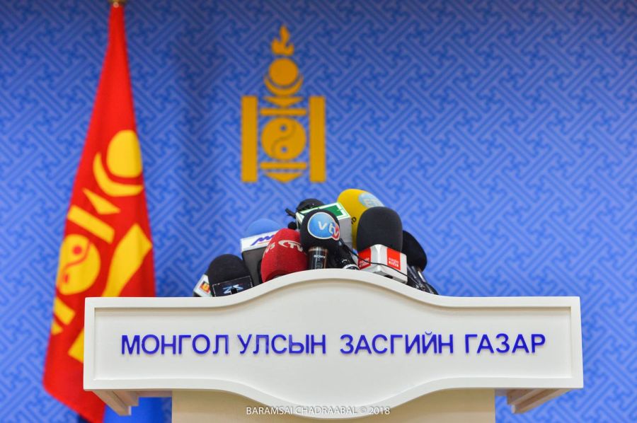 A podium with multiple microphones is in front of a backdrop featuring the emblem of Mongolia and text in Mongolian. A red flag with yellow elements is visible on the left.