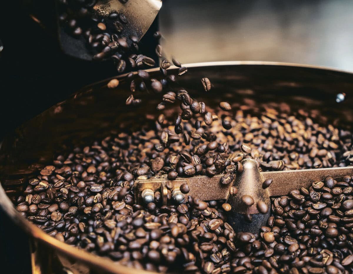 Металл шарах хүрд рүү цутгаж буй шарсан кофены шошны ойрын зураг. Шош нь бараан өнгөтэй, гялалзсан, бөмбөрийн дотоод хэсэг хэсэгчлэн харагдах бөгөөд шошыг хутгаж буй металл хутгагч гарыг харуулж байна. Энэ үзэгдэл нь цэцэглэн хөгжиж буй кофе бизнесийн мөн чанарыг харуулж байна. - Business.MN