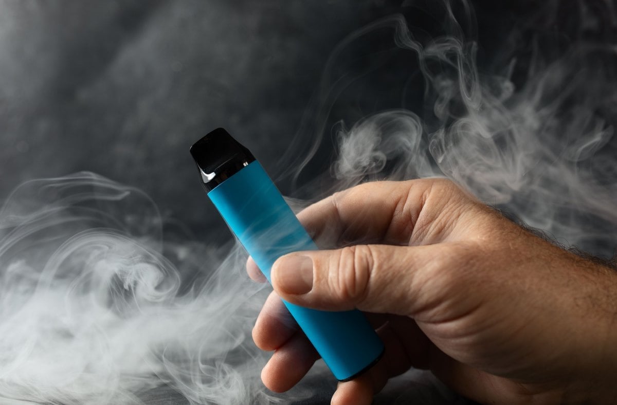 A hand holding a blue vape pen against a dark background, with smoke swirling around it.