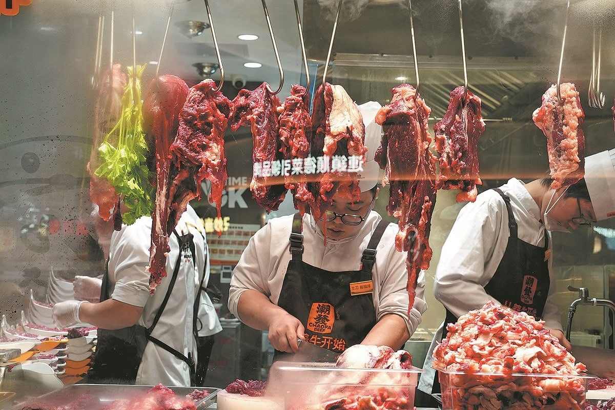 Гурван махны худалдаачин цагаан дүрэмт хувцас, малгай өмсөж, хөл хөдөлгөөн ихтэй зах, ресторанд улаан мах, навчит ногоо өлгөх шилэн дэлгэцийн ард чанартай мах бэлтгэж байна. - Business.MN