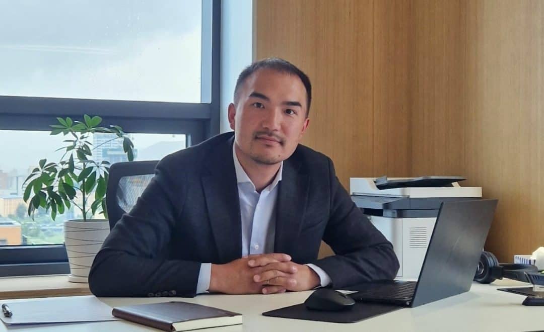 Ажил хэрэгч хувцастай эр оффисын ширээний ард сууж, урд нь зөөврийн компьютерээ онгойлгож байна. Ширээн дээр ваартай ургамал, принтер, дэвтэр бас байдаг. Арын арын том цонх нь байгалийн гэрлийг өгдөг. Энтрепренер нь төвийг сахисан илэрхийлэлтэйгээр камер руу шууд хардаг. - Business.MN