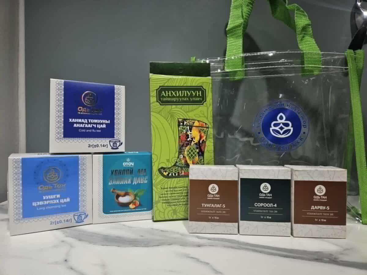 A collection of Mongolian herbal teas and skincare products displayed on a marble surface. Packaging features vibrant design elements and various text in Mongolian. A transparent green-handled bag is in the background.