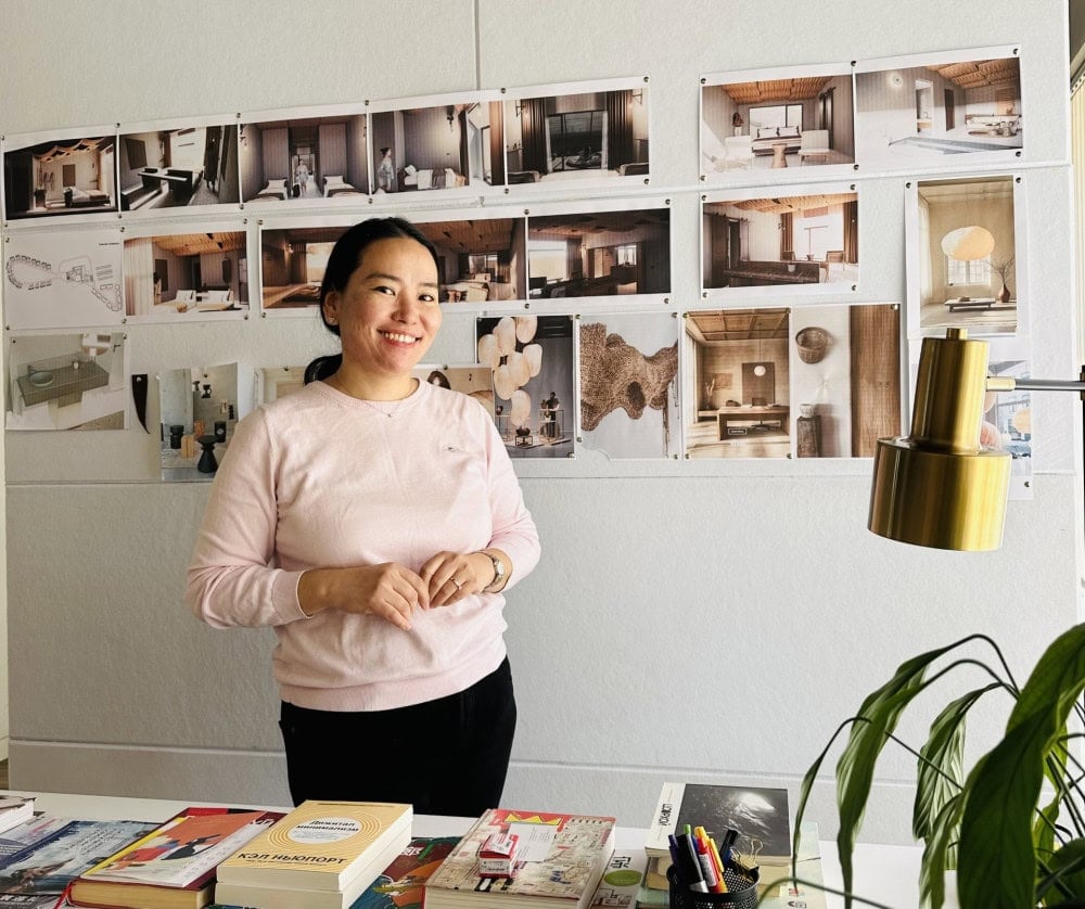 Өв соёлыг харуулсан интерьер дизайны гэрэл зургуудын хананы өмнө цайвар ягаан өнгийн цамц өмссөн эмэгтэй инээмсэглэн зогсож байна. Тэр ширээн дээр ном, цаас, дэнлүүтэй, урд талд нь ногоон ургамал байдаг. - Business.MN