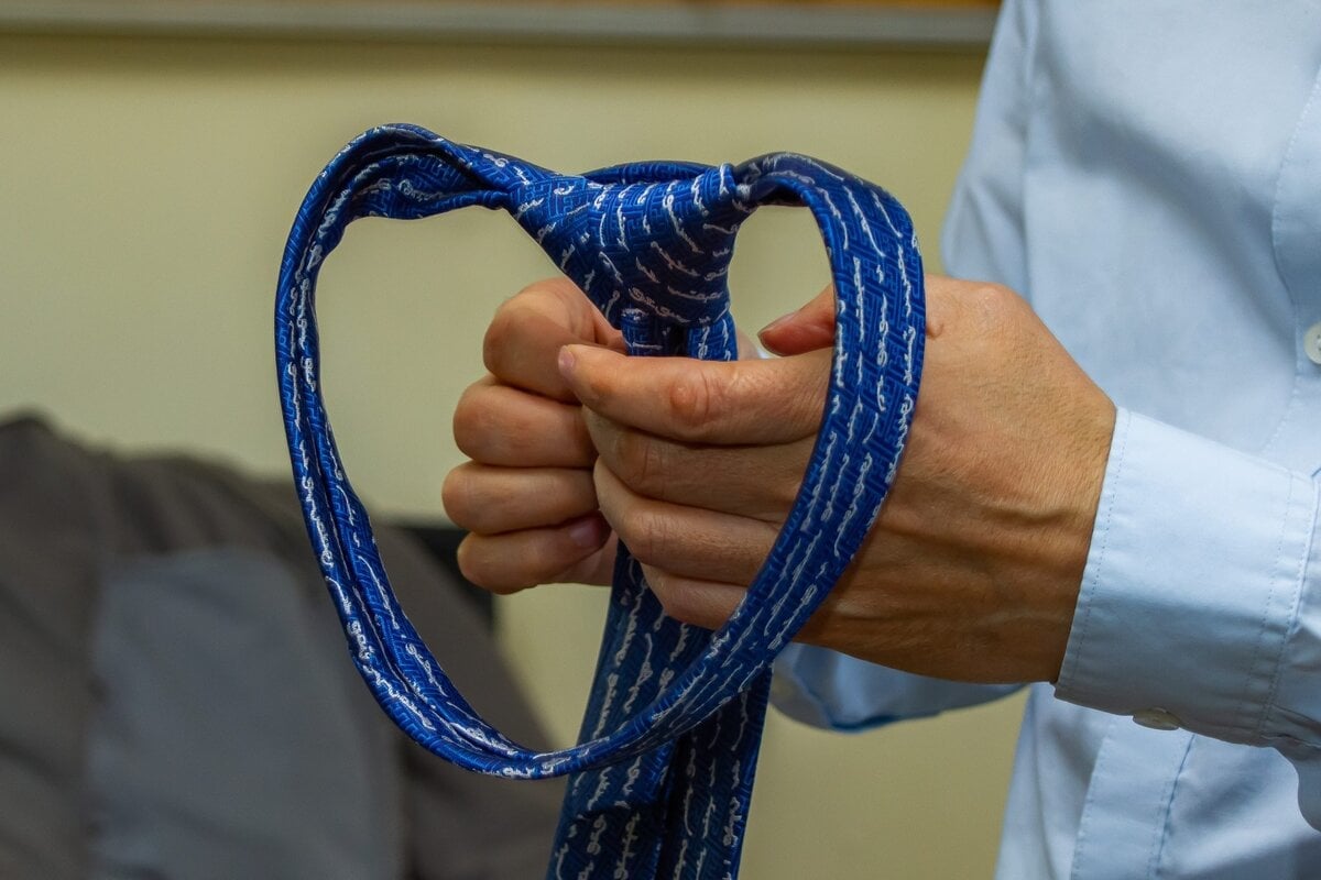 A person in a white shirt is tying a blue patterned necktie, holding the tie’s knot with both hands, with the wide end forming a heart-like shape.