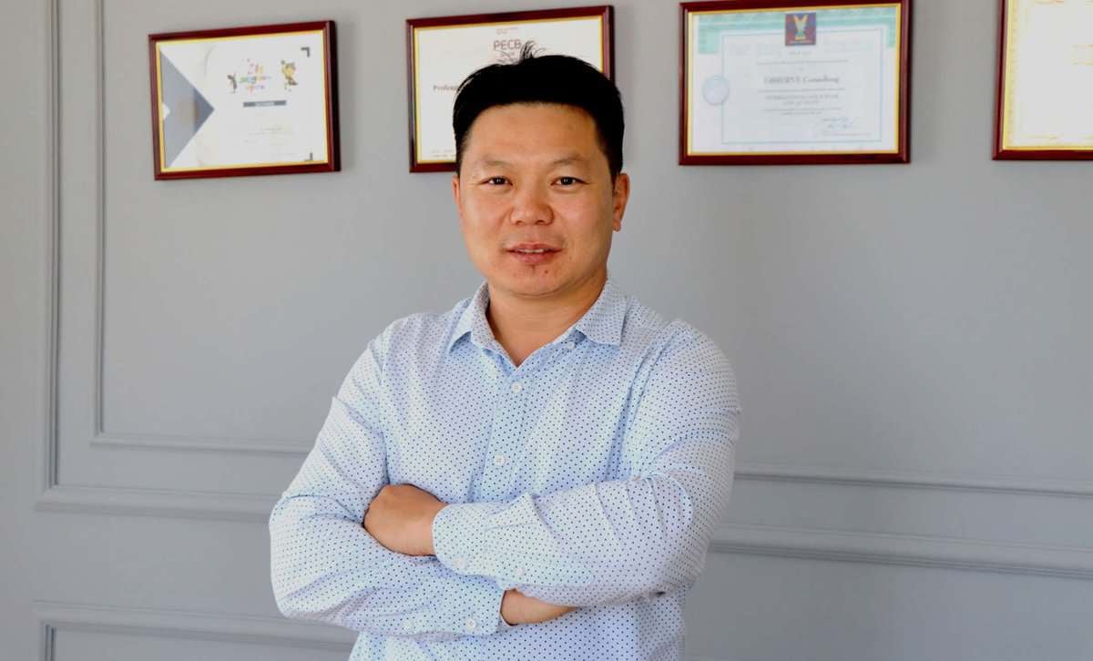 Цайвар цэнхэр цамцтай эр гараа зөрүүлэн зогсож, ардаа хананд наасан гэрчилгээнүүд нь энтрепренер болох түүний амжилттай аяллыг харуулж байна. - Business.MN